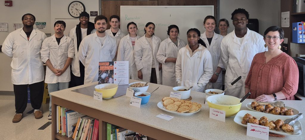 Student wearing White lab coats with their teacher standing behind a table with an array of food, some in bowls and some on plates.