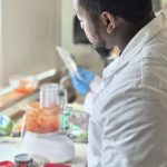 A student in a white lab watching food processor puree a red sauce on a counter with additional kitchen ingredients.