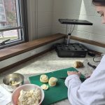 A student in a white lab coat chopping potato slices on green cutting board next to bowls of prepped cauliflower.