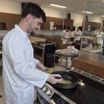 A student in a white lab coat sauteing onions in a pan on stove in a cooking lab.