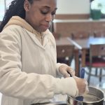 A Student in a white lab coat mixing ingredients in a saucepan with a blue spoon.