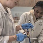 Two students in white chef jackets measuring and pouring a white liquid into a clear bowl