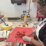 A woman cutting strawberries on a red cutting board with a measuring cup of strawberries on a scale, on a kitchen counter.