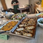 A display of cooked food in metal trays, some covered with tinfoil, and serving bowls and dishes with serving spoons on a table.