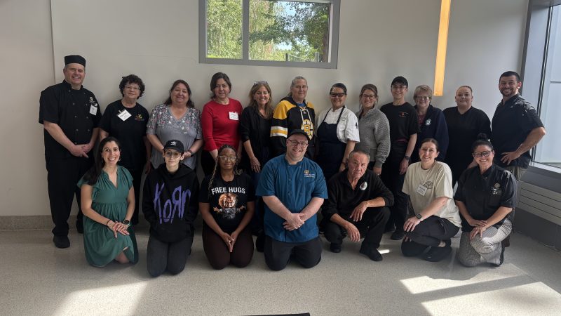 Chefs, staff, and participants pose for a group photo against a white wall with a large window, with some standing in the back row and others kneeling in the front, with a black rug in front of them.