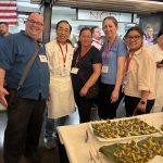 A group of 5 people posing for a photo near other people, wearing red lanyards in front of a table with two white platters with green leaf sheets on it and appetizers that are white with a brown topping and green garnish and toothpicks.