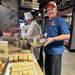 A Chef holding a metal bowl looking at the camera with many sandwiches with yellow bread.