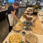 A woman in a white apron in a large room with tables, leaning over one table arranging a display of dishes on a variety of plates.