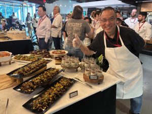 A Man in a white apron learning towards a table with a thumbs up in a room with other people. On the table is a display of food in different shapes and sizes of dishes.