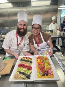 Two Chefs in aprons and chef hats in a kitchen standing behind a counter with two white plates, one with chopped avocado, raspberries, blueberries, and nuts. The other with green garnishes and fruit artfully displayed.