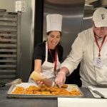 Two Chefs in a kitchen with the woman holding a bottle of brown sauce being drizzled on a sheet pan of brown circular pieces of food and man holds a piece of food.