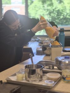 A woman holding a clear bag of yellow shredded cheese pouring it onto a tall scale on a white table with other kitchen appliances on it.