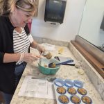 A woman wearing a striped shirt and black sweatshirt using a measuring cup to measure light brown muffin mix with yellow cheese and green jalapeno pieces in it, next to a cupcake pan filled with wrappers and muffin mix.