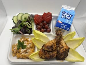 A school lunch tray with dairy pure milk, red grapes and cut strawberries, a salad with cucumbers, and ziti noodles in an orange sauce topped with cheese and cilantro, accompanied with 2 brown seasoned drumsticks on top of 6 endive leaves.