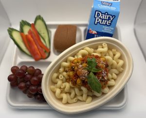 A bowl of pasta with a red sauce containing chickpeas, topped with basil next to red grapes, pieces of cucumbers and red peppers, a bread roll, and dairy pure milk, all on a lunch tray.