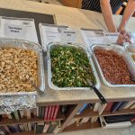 A counter with a display of cooked dishes in tinfoil pans with labels of the dishes in them.