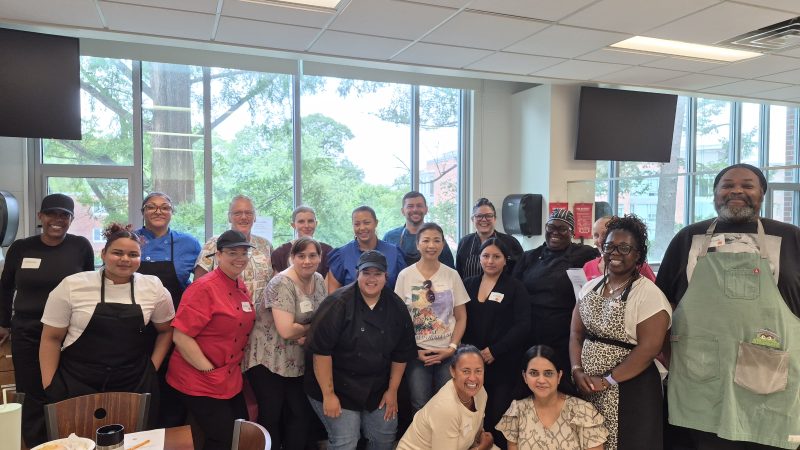 Group of chefs and staff, some wearing aprons and chef coats, standing and kneeling behind a table for a group photo in a kitchen with large windows.