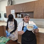 A woman in an apron standing next to another woman and a table in front of cabinets and counter holding a white bowl filed with brown seasoning.
