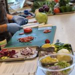 A persion wearing blue gloves standing in front of A counter with a green cutting board with different types of cuts of red tomatoes, and other bowls and pans showing cuts and scraps of onions, pineapples, jalapenos and other produce, with a variety of fruits and vegetables in the background.