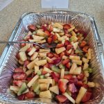A tinfoil pan on a tan counter with cut pieces of pineapple, strawberries, and kiwi with a serving spoon and a Small sign saying Tropical Fruit Salad