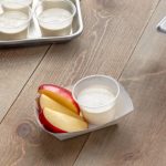 Three different serving kitchen items-a cookie sheet, a silver deep pan, and a small white paper serving container with small plastic portion cups filled with a white sauce and on the side red skin apple slices and whole red apple slices on a light brown wooden surface.