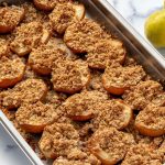 Close-up of baked pear halves topped with a golden brown oat crumble, arranged on a metal baking tray. Additional whole pears are visible beside the tray in the background.