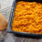A black plastic rectangular serving container filled with a orange mashed squash ontop of a silver metal wire rack on a grey wood service next to a tan/yellow colored squash and a circular dish with clear liquid in it.