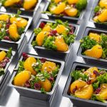 Eight black square containers on a metal tray, each holding a salad made of dark?green kale, bright?orange mandarin slices, deep?red dried cranberries, and tan sunflower seeds