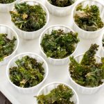 Close-up of crispy dark green kale crisps with lightly browned edges, served in small white cups arranged on a white tray.