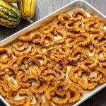A silver metal rectangular baking pan with white parchment paper under golden orange baked half cicular sliced pieces squash with green lines on skin. The Pan is ontop of a dark brown woooden surface with yellow with green stripes vegetables in the corner.