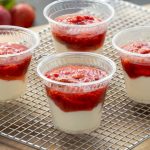 A silver wire tray ontop of light brown service with 4 clear small serivng cups, inside the there is a while liquid filling them halfway with a thick red sauce with strawberry pieces on the top with a blurred strawberry bunch in the background.