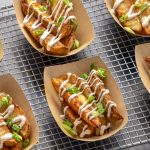 Six light brown paper trays filled with golden-brown roasted potato wedges are arranged on a metal wire rack. The wedge-cut potatoes are drizzled with a white creamy sauce and garnished with sliced green onions.