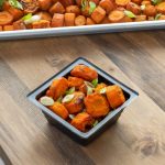 Close-up of roasted orange carrot slices coated in sauce and garnished with sliced green onions, served in a small black bowl on a wooden table, with a tray of additional roasted carrots in the background.