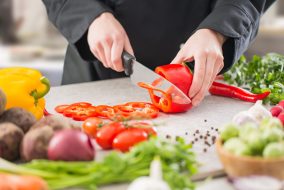 Cutting red pepper with a knife on a cutting board surrounded by other raw vegetables