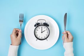 Hands holding knife and fork above alarm clock on white plate on blue background.