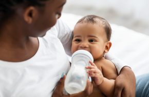 Childcare Concept. Closeup portrait of happy African American woman holding baby bottle and feeding her cute little black with milk at home. Nutrition, Love, Care And Family, Babysitting