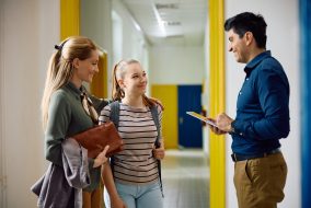 Happy teacher talks to high school student and her mother in hallway at school.